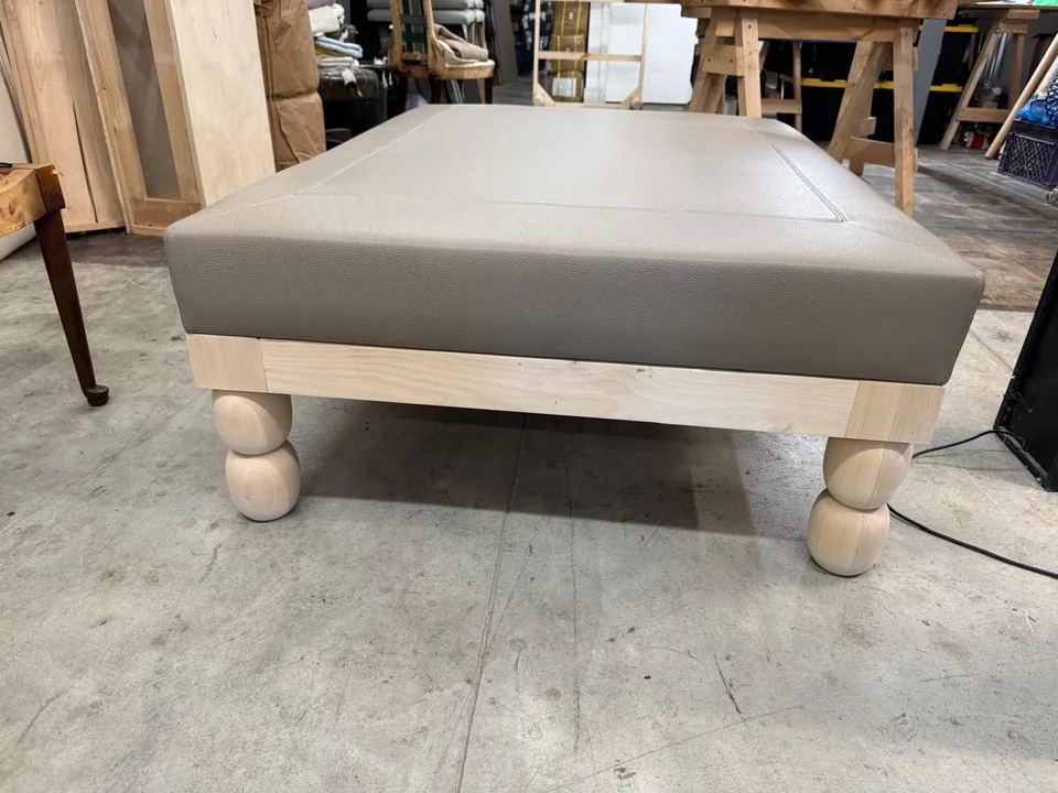 Front view of a taupe upholstered ottoman on a pale wooden frame